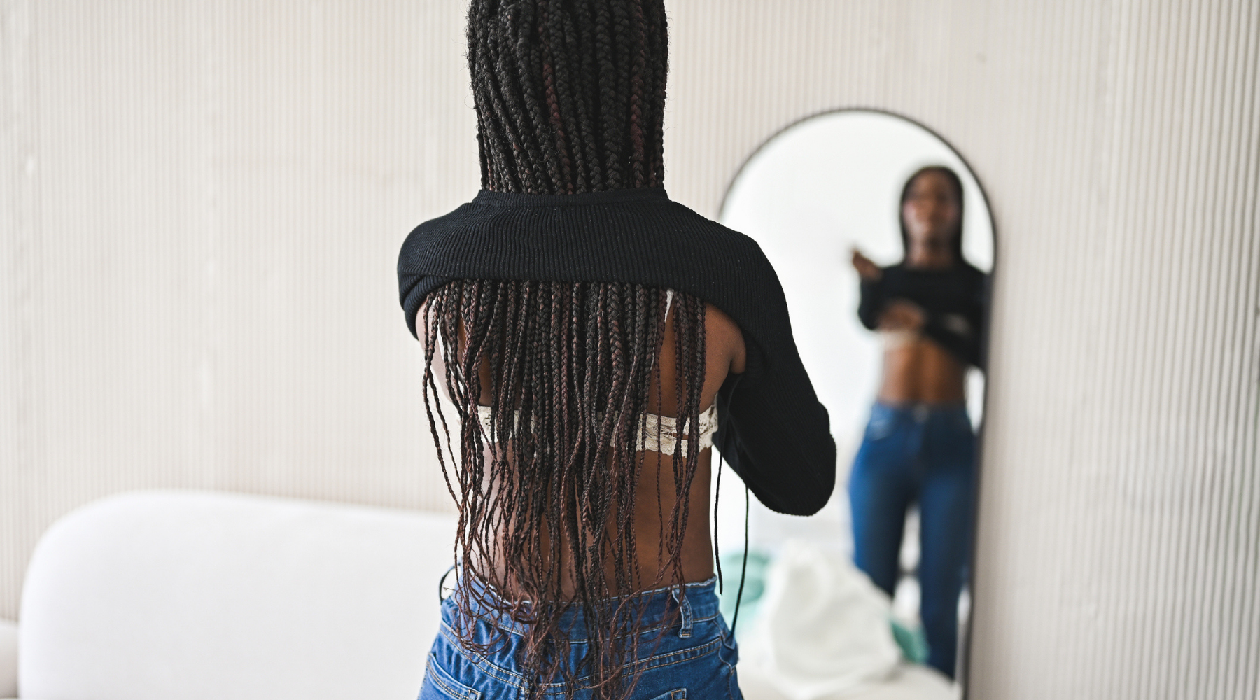 A women with braids stands in front of a mirror inspecting her stomach.